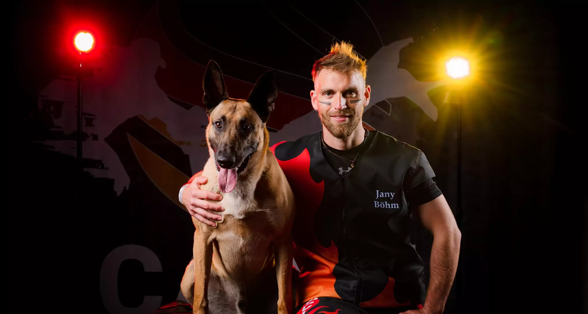 Jany Böhm sitzt mit schwarzem CACIT-Germany-Wettkampfshirt und schwarz-roter Gesichtsbemalung neben seinem Belgischen Malinois vor dem dramatisch beleuchteten CACIT-Germany-Backdrop.